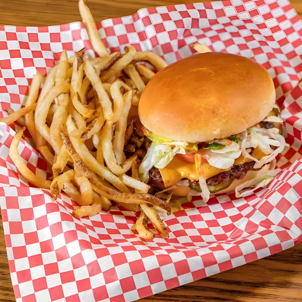 A Burger MOOvment cheeseburger with lettuce, tomato, and pickles, served with fries in a red and white checkered basket.